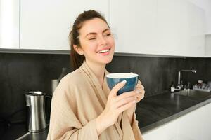 portrait de content Jeune femme départs sa Matin avec agresser de café, en buvant thé de tasse, permanent dans le cuisine, souriant gaiement photo