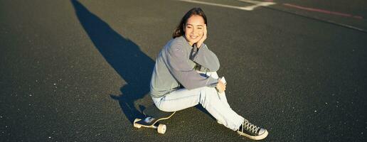 portrait de asiatique femme séance sur planche à roulette, patinage sur sa croiseur longboard, en utilisant téléphone intelligent app photo