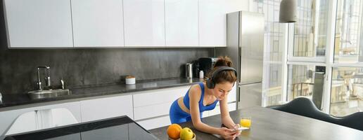 portrait de brunette femme dans tenue de sport, en utilisant téléphone intelligent, permanent dans cuisine, en buvant Frais Orange jus, en train de regarder faire des exercices Gym tutoriels photo