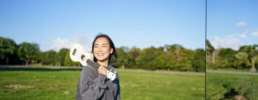 Jeune branché fille, voyageur en portant sa ukulélé, en jouant en plein air dans parc et souriant photo