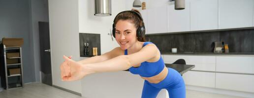 portrait de beau, en forme femme dans bleu leggings et soutien-gorge de sport, élongation sa bras, Faire aptitude entraînement, aérobie des exercices à maison, écoute la musique dans sans fil écouteurs photo