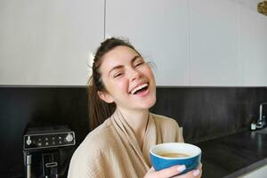 portrait de beau Jeune femme, en buvant café dans le cuisine, profiter sa Matin routine et souriant à le caméra photo