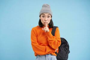 asiatique fille spectacles Chut, chut geste, met doigt plus de lèvres, garder silencieux signe, des stands plus de bleu Contexte avec sac à dos et porte chapeau photo