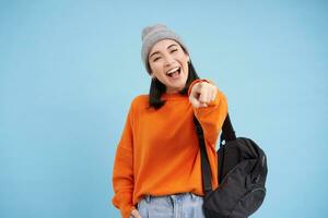 ses vous bravo. content souriant asiatique femme dans chapeau, points doigt à caméra, choisir, attrayant personnes, félicitant, permanent plus de bleu Contexte photo