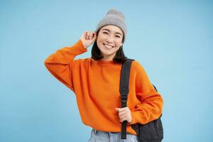 mignonne souriant asiatique fille met sur chaud chapeau à aller dehors, des promenades avec sac à dos dans Orange chandail, bleu Contexte photo