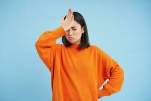 épique échouer. déçu asiatique femme claques sa front avec dérangé affronter, oublié qch, agacé, des stands plus de bleu Contexte dans Orange sweat-shirt photo