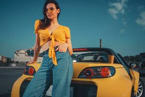 fille dans bleu jeans, Orange Haut et des lunettes de soleil est posant penché sur Jaune voiture cabriolet avec papier tasse de boisson sur tronc à parking parcelle. copie espace photo