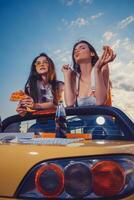 deux charmant femelles en mangeant Pizza tandis que posant dans Jaune voiture cabriolet avec un soda dans verre bouteille sur ses tronc. vite aliments. proche en haut, copie espace photo