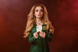 frisé blond fille dans vert élégant robe et bijoux. montrant deux en jouant cartes, posant sur coloré enfumé Contexte. poker, casino. fermer photo