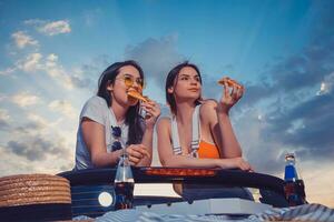 deux charmant femmes en mangeant pizza, posant dans voiture cabrio avec chapeau et un soda dans verre bouteilles sur ses tronc. vite aliments. été ensoleillé journée. proche en haut, copie espace photo