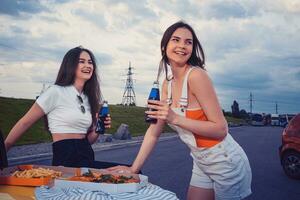 Jeune Dames sont souriant, profiter un soda dans verre bouteilles, posant penché sur tronc de Jaune voiture avec Pizza et français frites sur il. proche en haut, moquer en haut photo