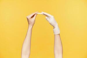 côté vue de Jeune féminin main tirant en dehors caoutchouc gant de autre main tandis que étant isolé plus de Orange Contexte. Humain mains et faire des gestes concept photo