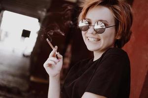 Fille aux cheveux roux courts et lunettes de soleil miroir fumant une cigarette