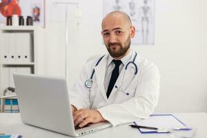 portrait de spécialiste médecin homme à la recherche dans caméra travail dans réunion conférence pièce en cours d'analyse maladie compétence en utilisant portable ordinateur. médecin prescrire pilules des médicaments soins de santé traitement photo