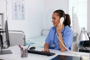 médical infirmière parlant avec patient sur le téléphone à propos diagnostic. santé se soucier médecin séance à bureau en utilisant ordinateur dans moderne clinique à la recherche à moniteur. photo