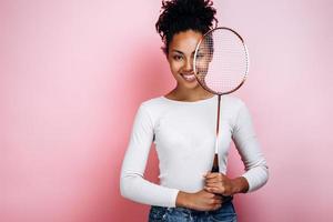 belle fille souriante se dresse sur un fond de mur rose, se couvre le visage avec une raquette photo