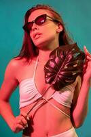 fille dans blanc bikini et des lunettes de soleil posant avec monstera feuille sur bleu Contexte photo