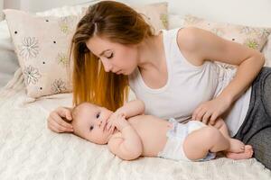 mère et enfant sur une blanc lit. maman et bébé fille dans couche en jouant dans ensoleillé chambre à coucher. photo