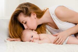 mère et enfant sur une blanc lit. maman et bébé fille dans couche en jouant dans ensoleillé chambre à coucher. photo