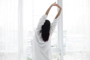 femme asiatique belle jeune femme souriante assise sur le lit et s'étirant le matin dans la chambre après s'être réveillée dans son lit complètement reposée et ouvrir les rideaux le matin pour prendre l'air frais. photo