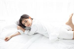 femme asiatique belle jeune femme souriante assise sur le lit et s'étirant le matin dans la chambre après s'être réveillée dans son lit complètement reposée et ouvrir les rideaux le matin pour prendre l'air frais. photo