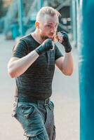 rue combattant dans noir vêtements et des pansements sur le poignet boxe dans perforation sac en plein air. Jeune homme Faire boîte formation et pratiquant le sien poinçons à le à l'extérieur Gym photo