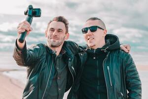 deux Beau souriant copains fabrication selfie en utilisant action caméra avec cardan stabilisateur à le plage. jeune Hommes dans noir vêtements ayant amusement par fabrication Photos
