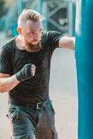 rue combattant dans noir vêtements et des pansements sur le poignet boxe dans perforation sac en plein air. Jeune homme Faire boîte formation et pratiquant le sien poinçons à le à l'extérieur Gym photo