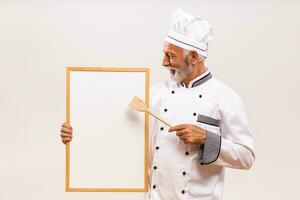 portrait de Sénior chef montrant avec en bois cuillère tableau blanc sur gris Contexte. photo