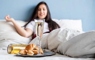 Jeune femme brune assise éveillée dans le lit avec des ballons en forme de coeur rouge et des décorations buvant du champagne en mangeant des croissants
