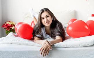 jeune femme brune heureuse pose dans le lit avec des ballons en forme de coeur rouge photo