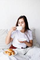 jeune femme brune assise éveillée dans le lit avec des ballons en forme de coeur rouge et des décorations buvant du café mangeant des croissants photo