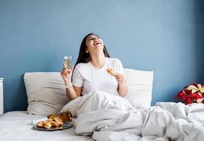 Jeune femme brune assise éveillée dans le lit avec des ballons en forme de coeur rouge et des décorations buvant du champagne en mangeant des croissants photo