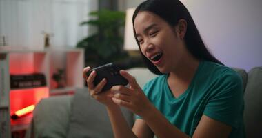 portrait de Jeune asiatique femme jouit en jouant en ligne esport Jeux sur téléphone intelligent séance sur canapé dans le vivant pièce à maison, joueur mode de vie concept, joueur mode de vie concept. photo