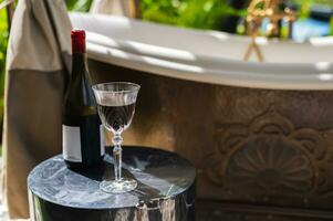 une verre de rouge du vin avec bouteille mettre sur marbre table à côté de Extérieur baignoire à prendre plaisir et relaxant moment. photo