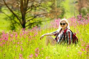 content aventurier femelle des stands sur le vert Montagne pente parmi floraison rose rhododendrons et à la recherche dans le distance. épique Voyage dans le montagnes. large angle. photo