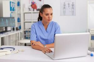 médical infirmière travail sur portable dans hôpital Bureau portant bleu uniforme. santé se soucier praticien séance à bureau en utilisant ordinateur dans moderne clinique à la recherche à moniteur, médicament. photo