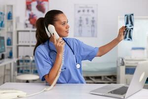 médical infirmière en portant patient radiographie dans hôpital Bureau tandis que parlant avec médecin sur téléphone. santé se soucier médecin séance à bureau en utilisant ordinateur dans moderne clinique à la recherche à moniteur. photo