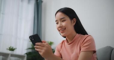 portrait de content Jeune asiatique femme se détendre sur confortable canapé à Accueil achats en ligne sur téléphone intelligent, souriant fille utilisation cellule téléphone bavardage, Feuilleter sans fil l'Internet sur gadget photo