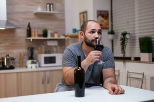 caucasien homme en portant verre de du vin séance dans cuisine à maison. déprimé la personne en buvant alcool, biberonner, alcoolique boisson seul tandis que sentiment ivre avec bouteille de de l'alcool photo