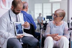 spécialiste médecin montrant X rayon à personnes âgées femme dans fauteuil roulant communicant tester résultat en utilisant tablette ordinateur. médecin expliquant traitement maladie pendant récupération tandis que séance dans hôpital zone photo