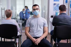 portrait de souriant Jeune patient à la recherche sur caméra séance sur chaise dans attendre pièce de hôpital clinique respectant social distanciation tandis que médecin travail dans Contexte. assistant dactylographie sur ordinateur photo