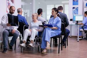 médecin avec stéthoscope parlant et montrer du doigt à radiographie de désactivée vieux femme dans fauteuil roulant, expliquer traitement séance dans hôpital attendre zone. paralysé patient discuter avec médical Personnel photo