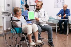 médecin montrer du doigt sur tablette PC avec vert écran à la recherche à handicapé Sénior femme. médical praticien en utilisant dispositif avec chrominance clé pendant consultation de invalide Sénior femme. photo