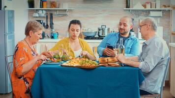 homme en utilisant téléphone pendant dîner montrant certains des photos à sa mère. multi génération, quatre personnes, deux content des couples parlant et en mangeant pendant une gourmet repas, profiter temps à maison.