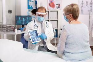 médecin en portant tablette PC avec radiographie de Sénior femme dans hôpital dans le cours de COVID-19 [feminine. médical médecin spécialiste pendant coronavirus déclenchement discuter avec patient. photo