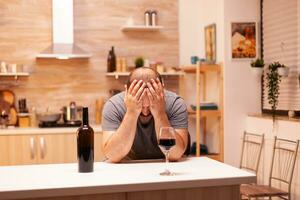 homme ayant difficulté avec de l'alcool pendant la vie crise avec une bouteille de du vin dans cuisine séance à tableau. malheureux la personne maladie et anxiété sentiment épuisé avec ayant alcoolisme problèmes. photo