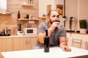 dérangé Jeune homme en buvant seul une bouteille de rouge du vin séance à table dans cuisine. malheureux la personne maladie et anxiété sentiment épuisé avec ayant alcoolisme problèmes. photo