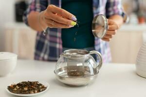 proche en haut de verser aromatique herbes dans théière à faire thé dans Matin pour petit déjeuner. en train de préparer thé dans le matin, dans une moderne cuisine séance près le tableau. en mettant avec mains, en bonne santé à base de plantes dans pot. photo