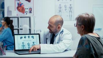 Jeune Masculin médecin expliquant Comment à éviter des os problèmes à personnes âgées retraité vieux femme. radiologie et radiographie dans moderne privé hôpital ou clinique avec médical Personnel en marchant dans arrière-plan, infirmière fonctionnement, soins de santé système. photo